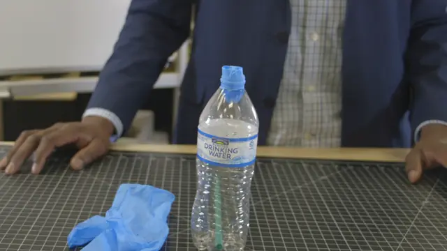 A water bottle on a cutting mat, the finger of a rubber glove pulled down into its neck, the glove beside it.