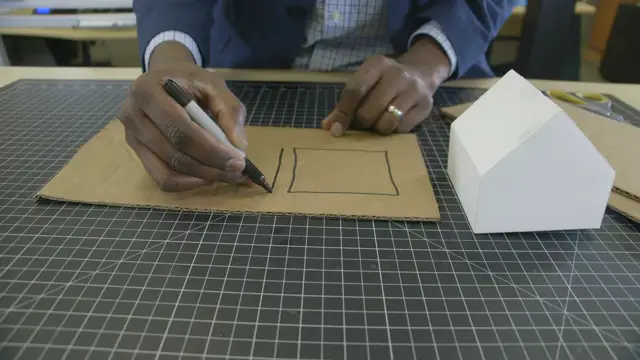 Hands drawing a wall piece on flat cardboard with a marker, a white model house beside it.