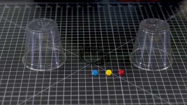 Two plastic cups, a coil of string and three push pins on a cutting mat.