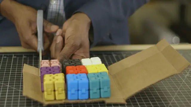 Hands trimming a folded cardboard stand with scissors, colorful toy cubes loaded inside it.