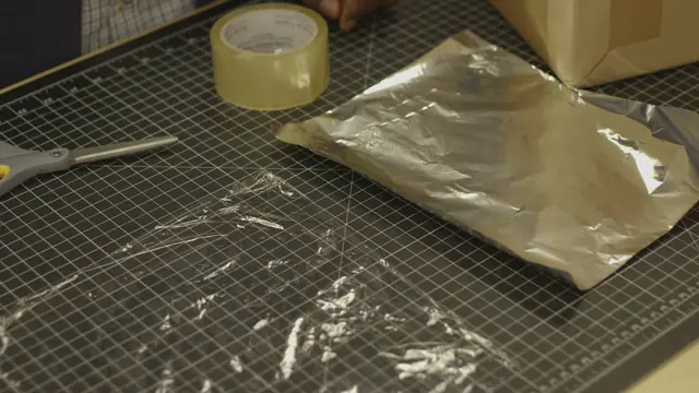 A roll of tape, scissors, a foil-covered sheet of cardboard and crumpled plastic wrap on a cutting mat.