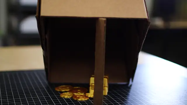 A cardboard box propped up on a stick over a pile of gold coins.